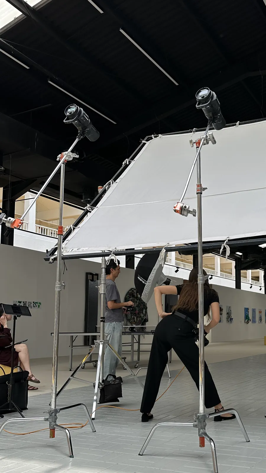 Behind the scenes on the Back to School 2025 shoot: two lights on tall stands over a large diffusion scrim, with the crew setting up in a school hall.