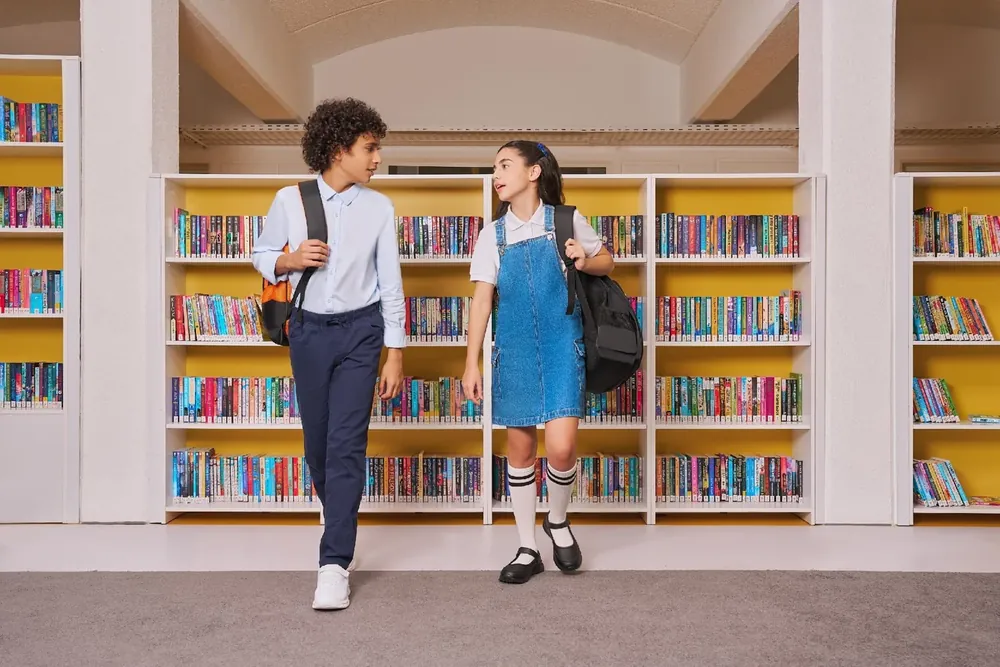 LC Waikiki Back to School 2025 campaign photograph: two students with school bags walking past library shelves.