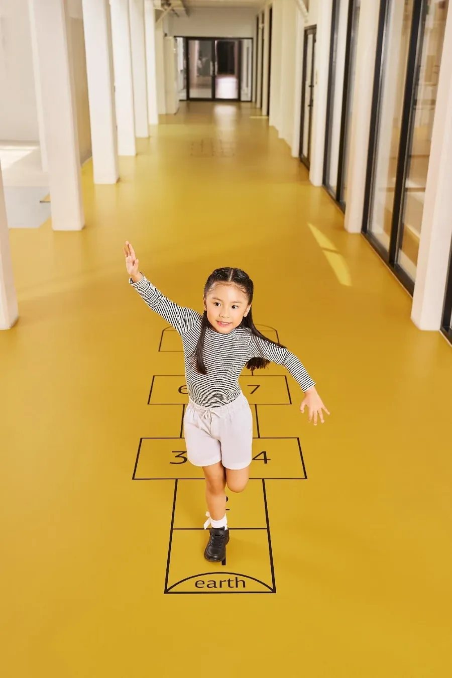Campaign photograph of a girl playing hopscotch in a yellow school corridor.