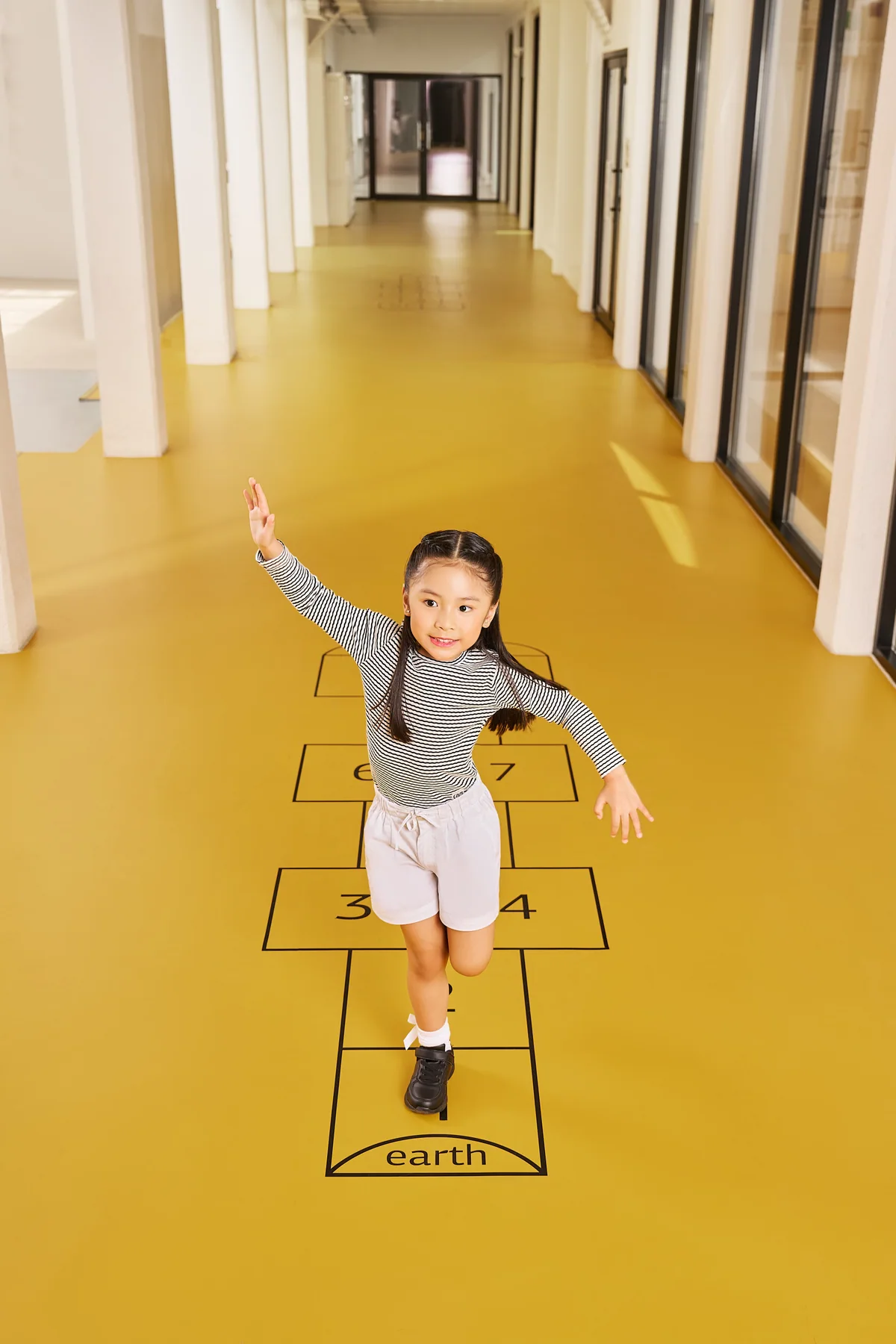 Campaign photograph of a girl playing hopscotch in a yellow school corridor.